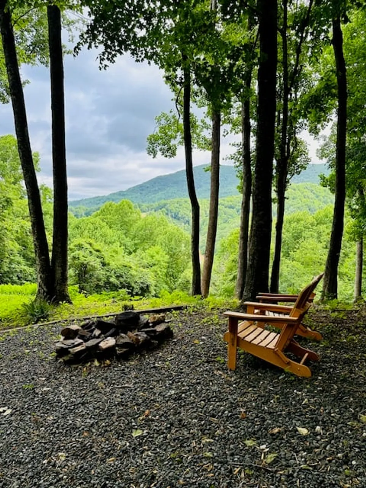 A rustic fire pit surrounded by wooden chairs looks out over a stunning mountain view, framed by trees. The setting is ideal for evening gatherings, storytelling, and stargazing under the open sky.