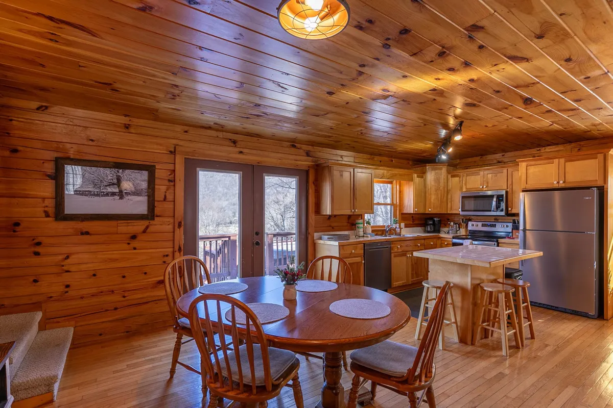 This image features a spacious wooden dining table set adjacent to kitchen cabinets, all bathed in natural light from large windows and complimented by a wooden ceiling. The setup is inviting and ideal for family meals or group gatherings, capturing the essence of communal dining.