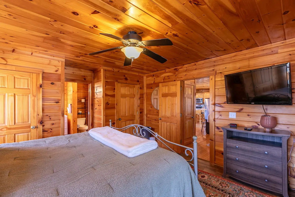 A comfortable bedroom featuring a well-appointed bed, a ceiling fan for comfort, a TV for evening entertainment, and warm wooden doors, all bathed in natural light.