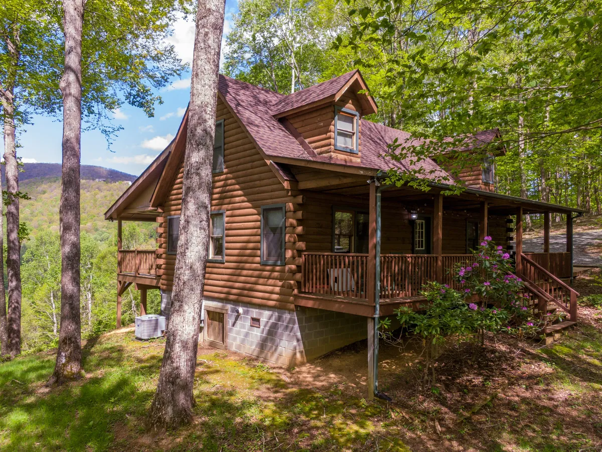 A log cabin sits nestled among trees, with a spacious front porch overlooking mountain scenery. The setting hints at immediate access to nature, with outdoor spaces ideal for preparing gear, relaxing, or launching into nearby adventures.