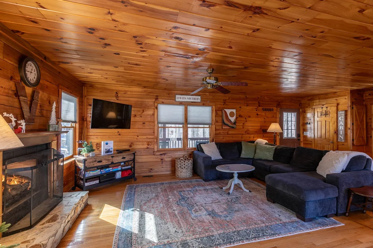 A cozy living room scene with a prominent stone fireplace, plush sofa, textured rug, and a Smart TV set against rustic wooden walls. Large windows invite natural light, and the seating arrangement encourages both relaxation and social interaction.