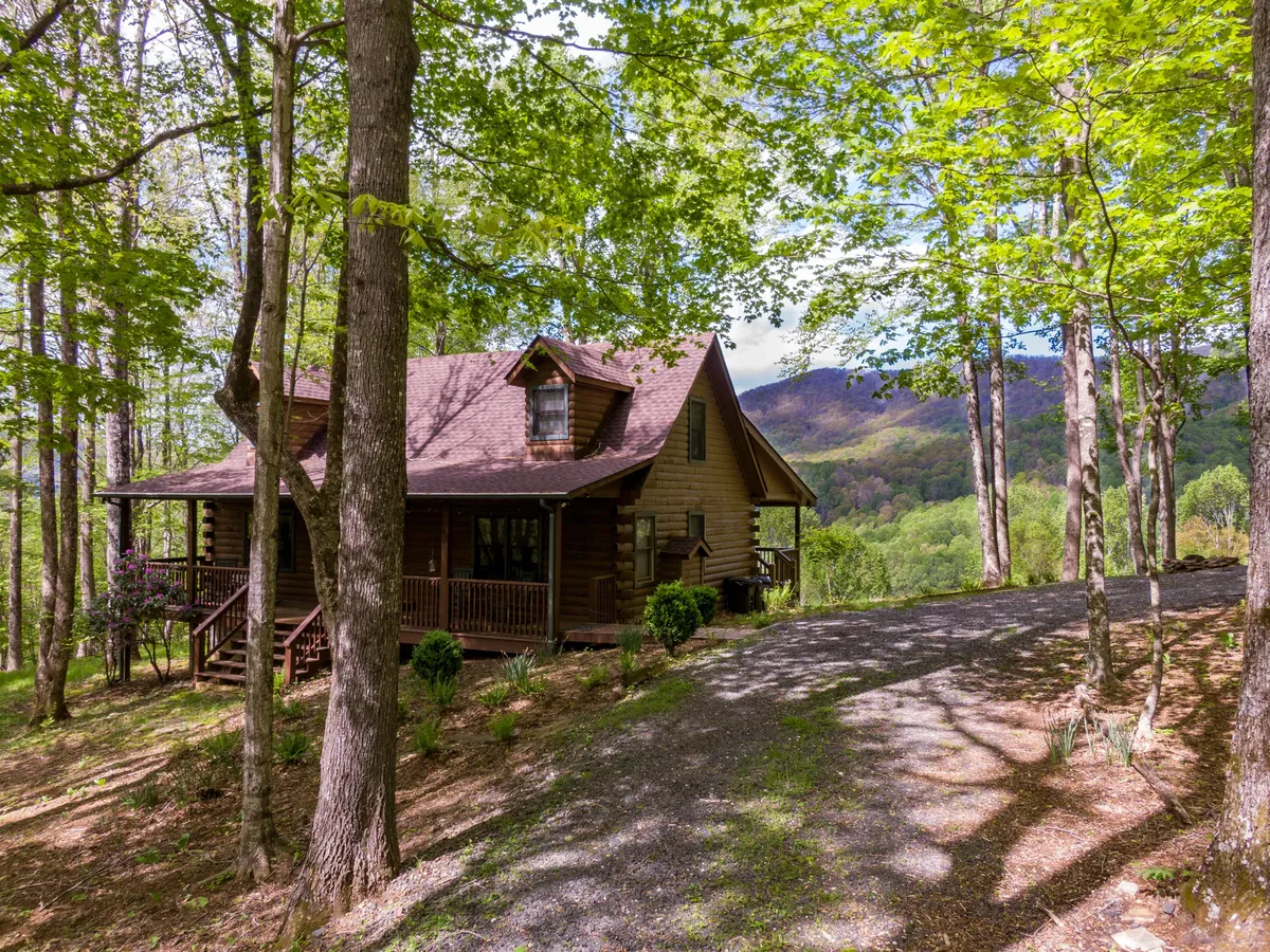 The image features a charming wooden house nestled among lush trees, with expansive views of the Blue Ridge mountains stretching into the distance. The scene exudes tranquility and natural beauty, with a spacious porch perfect for relaxing and soaking in sunsets or stargazing under the clear mountain sky.