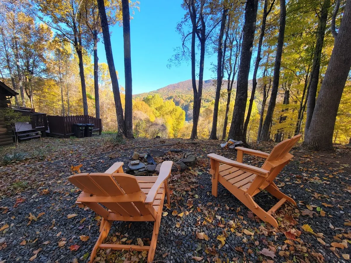 An exterior scene showing two wooden chairs arranged around a fire pit, set against a panoramic backdrop of mountains. The mood is serene and invites relaxation, conversation, and stargazing.
