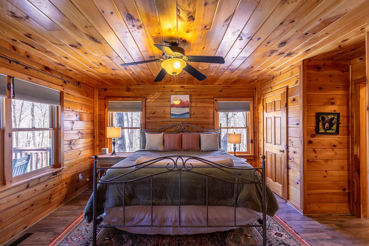 A cozy bedroom with a plush bed neatly made with fresh linens, flanked by windows that allow soft natural light to stream in. The room is accented by a nightstand with lamps and tasteful artwork, creating a serene and restful atmosphere.