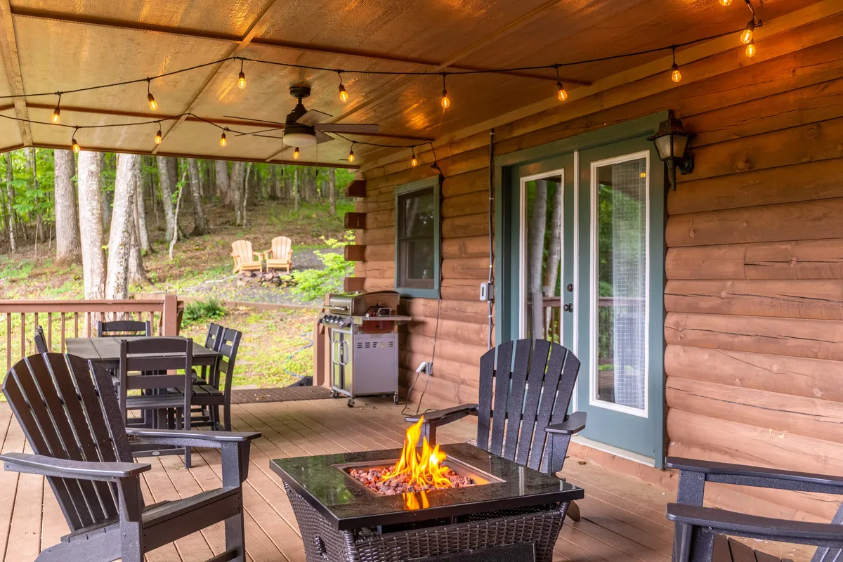 A cozy fire pit area set against the log cabin wall, surrounded by wooden chairs and illuminated by twinkling string lights. The space invites guests to gather, relax, and enjoy the outdoors—perfect for evening conversations or roasting marshmallows.