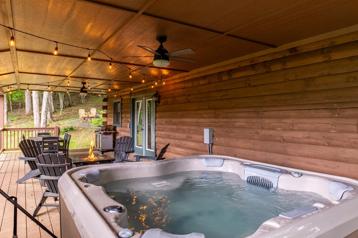 This exterior image captures a bubbling hot tub illuminated by string lights, with outdoor seating nearby and a fire pit in the background. The setting is surrounded by nature, contributing to an intimate and tranquil ambiance ideal for couples.
