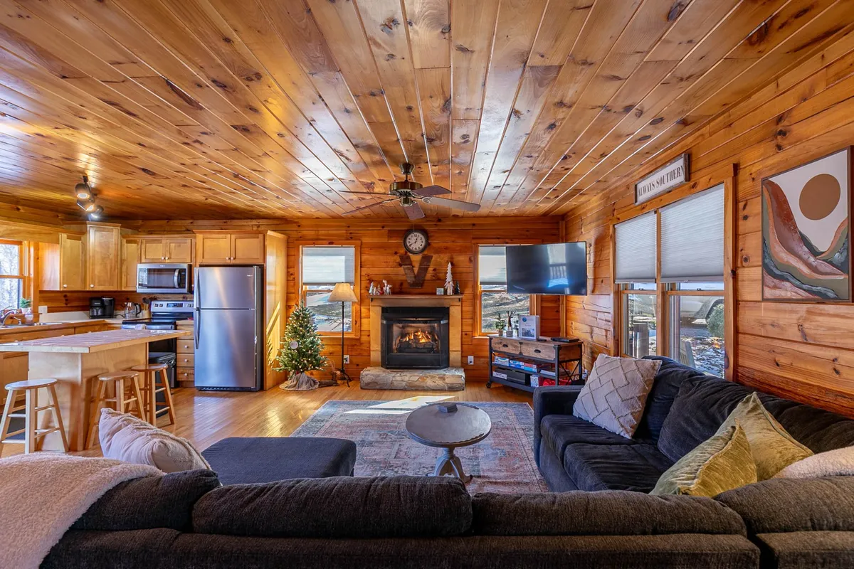 A warm, inviting living space featuring a central gas log fireplace surrounded by a comfortable sofa and coffee table. The living area blends seamlessly with a kitchen space in the background, creating an open, communal environment ideal for gatherings. Artwork adorns the wall, enhancing the homely ambiance.