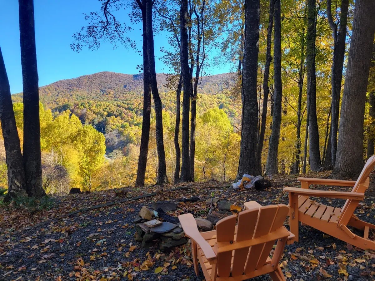 A tranquil fire pit area with wooden chairs overlooking a beautiful mountain view and vibrant autumn foliage. The scene suggests peaceful mornings, nature appreciation, and group storytelling sessions as the sun sets.